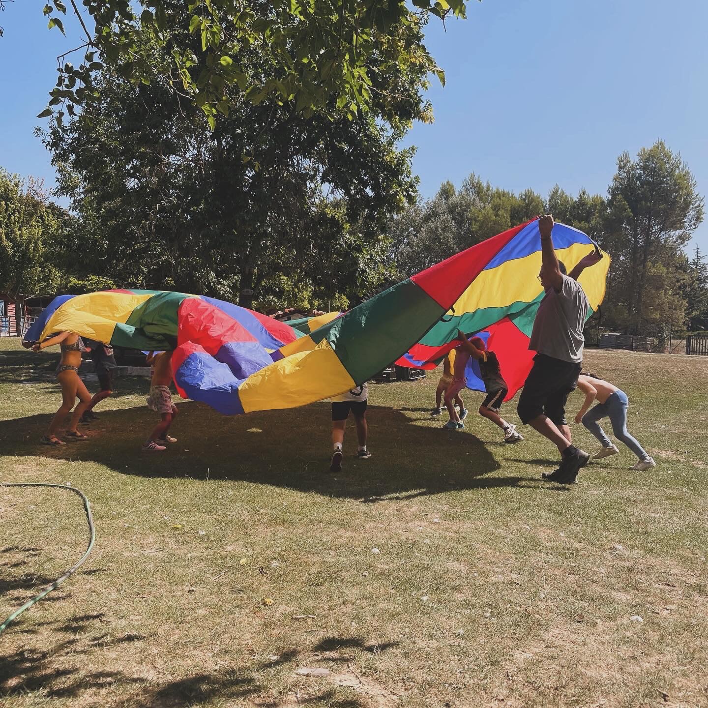 Niños y monitor participando en un juego cooperativo con paracaídas durante actividades extraescolares en la granja escuela Naturalia de Esquíroz